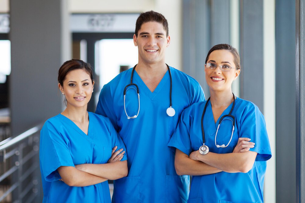 Operating room assistants smile at the camera. They are all wearing blue work clothes. Symbol for healthcare, advisory, care, hospital, merger acquisition, sale, consultant, consulting, mergers, acquisitions, M&A, company sale